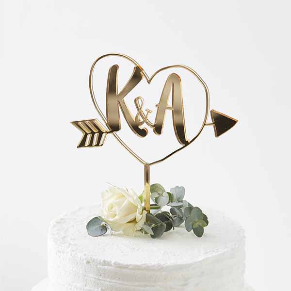 A gold acrylic cake topper is displayed on a white frosted cake. The topper features a heart shape surrounding the initials &quot;K&amp;A,&quot; with an arrow pointing outward. Nestled at the base of the topper is a small bouquet consisting of a white rose and eucalyptus leaves, adding a touch of elegance to the cake&