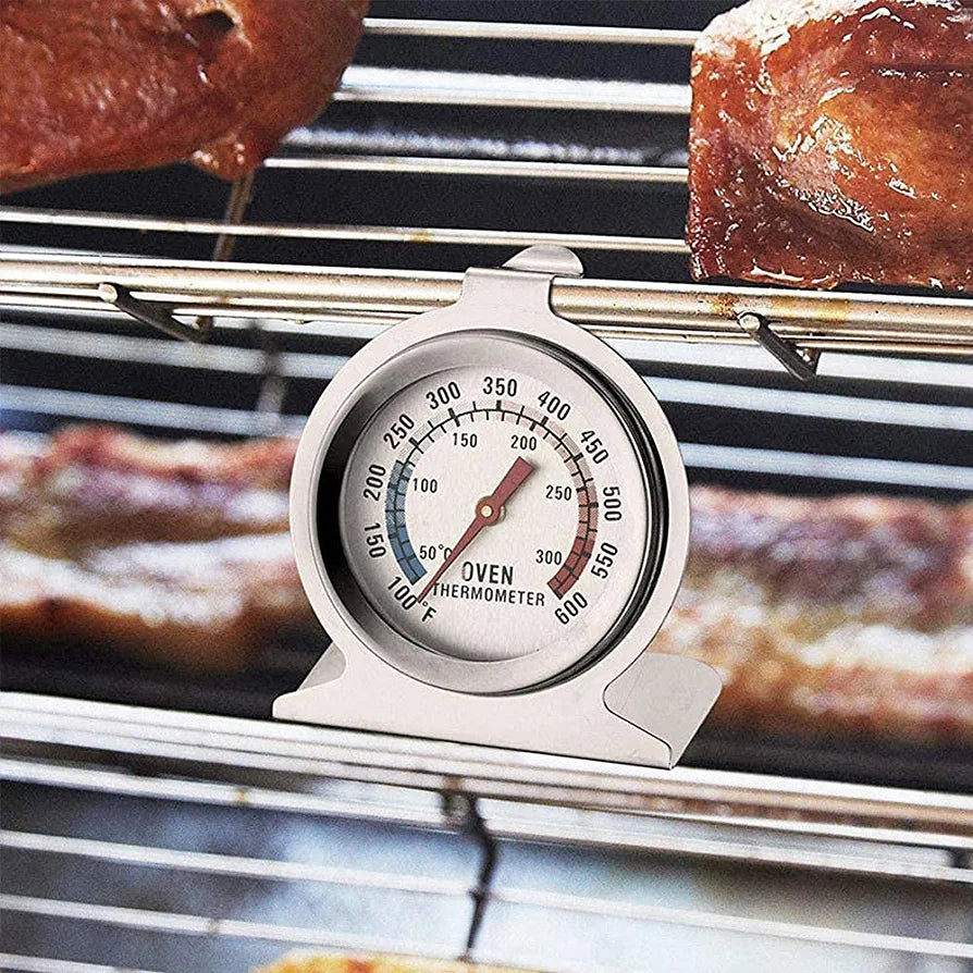 An oven thermometer is displayed on a metal rack inside an oven. The thermometer features a round dial with a white background, marked with temperature readings from 100°F to 600°F, and a bright red needle indicating the current temperature. The word "OVEN" is prominently displayed on the dial. In the background, there are baked goods, suggesting a cooking environment. The thermometer is designed to stand upright on the oven rack.