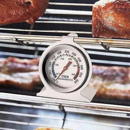 An oven thermometer is displayed on a metal rack inside an oven. The thermometer features a round dial with a white background, marked with temperature readings from 100°F to 600°F, and a bright red needle indicating the current temperature. The word "OVEN" is prominently displayed on the dial. In the background, there are baked goods, suggesting a cooking environment. The thermometer is designed to stand upright on the oven rack.