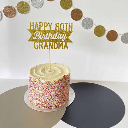 A round cake with a smooth vanilla frosting and colorful sprinkles on the sides sits on a silver cake board. Topped with a glittery gold cardstock cake topper that reads "HAPPY 80TH Birthday GRANDMA." In the background, there is a decorative garland made of circles in gold, silver, and brown. The table is covered with a mix of light and dark colored surfaces.