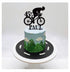 A decorated cake featuring a blue frosting top with mountain designs and clouds, a green grass-like texture at the base, and a black silhouette of a cyclist riding a bicycle on top, along with the number "45" and the name "PAUL" in black lettering. The cake sits on a round black base with white road markings.