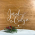 The image features an acrylic wedding cake topper in the shape of a heart. The topper displays the names "Jared" and "Cailyn" in stylish, elegant white script. Below the names, the date "02.27.2021" is included, accompanied by a small heart graphic. The topper is placed on a white cake, with a decorative arrangement of greenery and red berries underneath it. The background is a wooden surface, enhancing the overall rustic and romantic appearance.
