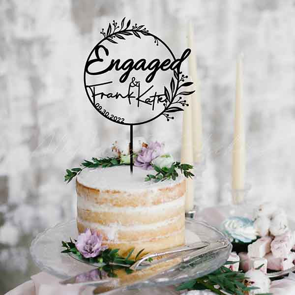 A simple layered cake displayed on a glass stand with a textured, white icing on top. Atop the cake, there is a black acrylic cake topper featuring the words "Engaged &amp; Frank Kate" surrounded by decorative leaf designs, and the date "09.30.2022" below. The cake is adorned with green leaves and small purple flowers. In the background, there are two tall, cream-colored candles and a softly blurred backdrop.
