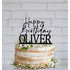 A round, multi-layered cake is decorated with a black acrylic cake topper that reads "Happy Birthday OLIVER" in a combination of script and bold font. The cake has a rustic appearance, with visible layers and light frosting. It is adorned with small white flowers and greenery around the base. The background features a simple, light-colored brick wall, enhancing the festive feel of the scene.