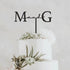 A three-tier white wedding cake decorated with greenery and succulents. The cake features a black personalized cake topper displaying the initials "M" and "G" connected by the word "and" in a cursive font. The background is soft and neutral, enhancing the elegance of the cake design.
