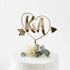 A gold acrylic cake topper is displayed on a white frosted cake. The topper features a heart shape surrounding the initials "K&A," with an arrow pointing outward. Nestled at the base of the topper is a small bouquet consisting of a white rose and eucalyptus leaves, adding a touch of elegance to the cake&