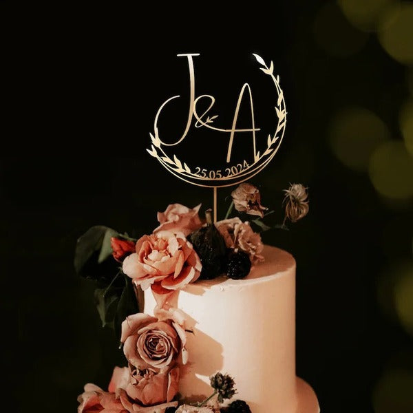 A wedding cake topper featuring the initials "J&amp;A" in a stylish font, surrounded by a circular floral design. Below the initials, the date "25.05.2024" is displayed. The cake is adorned with fresh roses in soft pink and cream hues, along with green foliage, creating an elegant and romantic presentation. The background appears dark, enhancing the visual focus on the cake and topper.