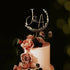A wedding cake topper featuring the initials "J&A" in a stylish font, surrounded by a circular floral design. Below the initials, the date "25.05.2024" is displayed. The cake is adorned with fresh roses in soft pink and cream hues, along with green foliage, creating an elegant and romantic presentation. The background appears dark, enhancing the visual focus on the cake and topper.
