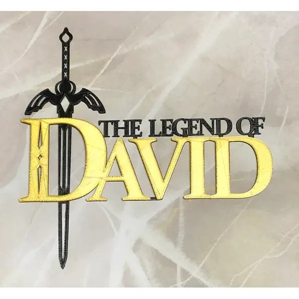 A cake topper features a decorative design with the words "THE LEGEND OF DAVID" in bold fonts. The word "DAVID" is notably larger and in gold, while the rest of the text is black. Above the text, there is a stylized sword graphic. The background appears to have a light, marbled texture.