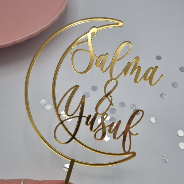 The image features a personalized cake topper designed for weddings. It is shaped like a crescent moon and made of gold-colored material. The topper has the names "Salma" and "Yusuf" elegantly scripted, with an ampersand (&amp;) between the names. The background includes a few scattered silver confetti pieces, and a soft pink plate is visible at the edge of the image.