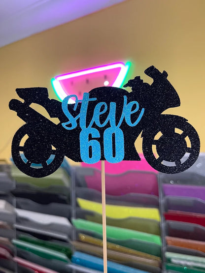 A cake topper made from black glitter cardstock features a motorcycle silhouette on either side. The text "Steve" is prominently displayed in a cursive blue font, with the number "60" in a bold blue font below it. The background shows colorful sheets of cardstock in various colors, and there is a soft glow of neon lights in the upper part of the image.