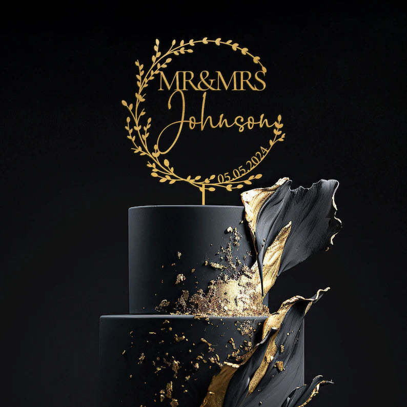 A black three-tier cake adorned with a gold-accented design. At the top, a personalized acrylic cake topper is displayed, featuring the text "MR &amp; MRS Johnson" in an elegant font, surrounded by decorative leaves, along with the date "05.05.2024." The background is dark, enhancing the cake&