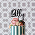 A personalized acrylic cake topper featuring the name "Billy" is placed atop a beautifully decorated cake. The cake has alternating dark blue and white vertical stripes and is adorned with colorful, delicate floral decorations in shades of pink, cream, and green. The background consists of a subtle, patterned design that adds an elegant touch to the overall presentation.
