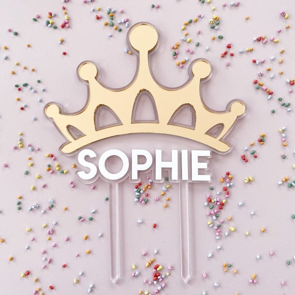 A decorative cake topper featuring a gold crown design with the name "SOPHIE" in bold white letters below. The topper has two clear support sticks for insertion into a cake. The background is a light color scattered with colorful, small confetti sprinkles.