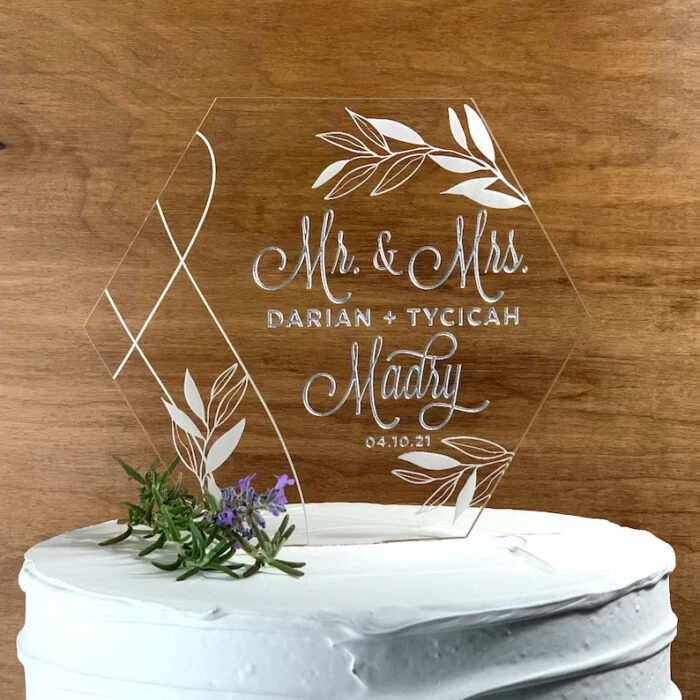 A hexagonal wedding cake topper made of clear acrylic, featuring elegant script text that reads "Mr. &amp; Mrs." followed by the names "Darian  Tyciciah Madry" and the date "04.10.21". The topper is adorned with leafy designs and placed on a white, textured cake topped with small greenery and purple flowers, set against a wooden background.
