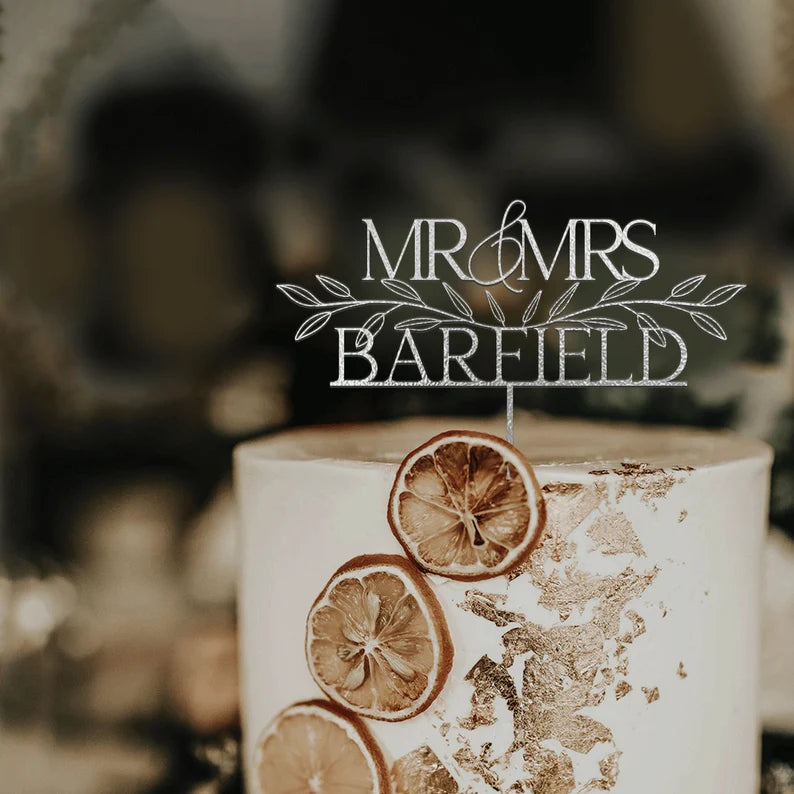 A white, cylindrical wedding cake topped with an acrylic cake topper. The topper features the words "MR &amp; MRS" above the name "BAREFIELD" decorated with leafy accents. The cake is adorned with dried orange slices and gold leaf accents on its surface. The background is softly blurred, highlighting the cake and topper.