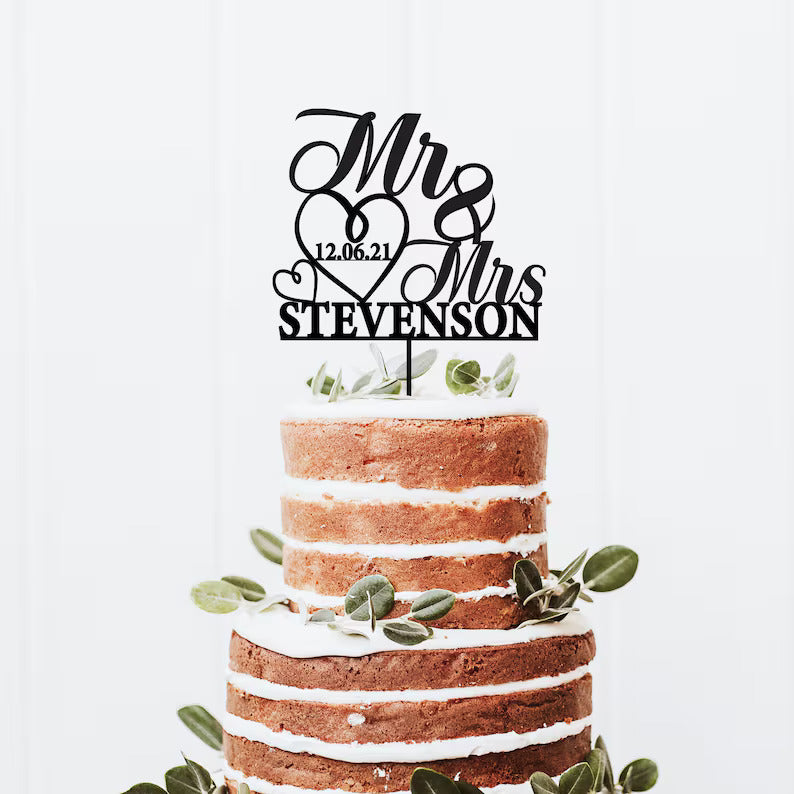 A three-tiered, semi-naked wedding cake with a rustic appearance features alternating layers of sponge and frosting. The top of the cake is adorned with a black cake topper that reads "Mr &amp; Mrs" along with the name "Stevenson" and the date "12.06.21." The cake is decorated with fresh greenery, adding a natural touch to the design. The background is a light-colored wall, enhancing the cake&