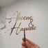 A hand is holding a personalized wedding cake topper made of gold-colored material. The topper features the names "Ahcene" and "Hananë" in elegant, cursive lettering, along with an ampersand symbol (&) between the names. The background is light-colored with decorative elements like stars and clouds, enhancing the celebratory theme of the item.