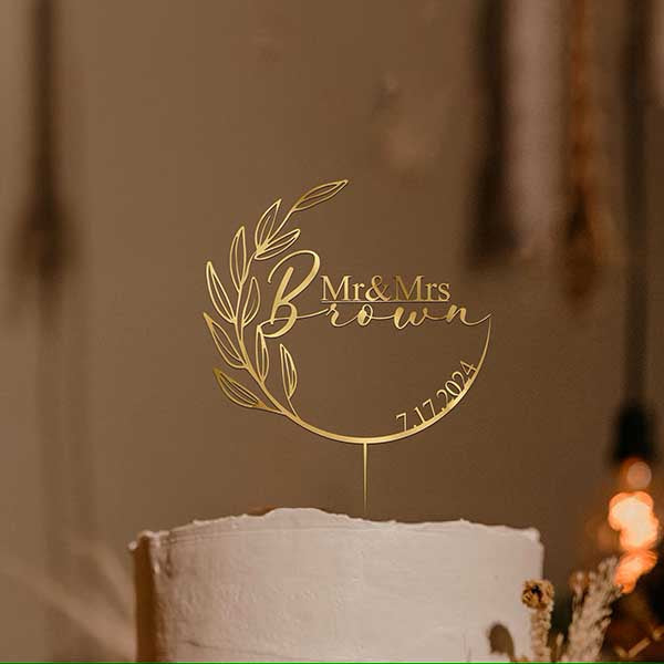 A golden acrylic cake topper featuring the text "Mr &amp; Mrs Brown" with decorative leaves and the date "7.17.2024." The topper is placed on a white wedding cake in a softly lit setting.