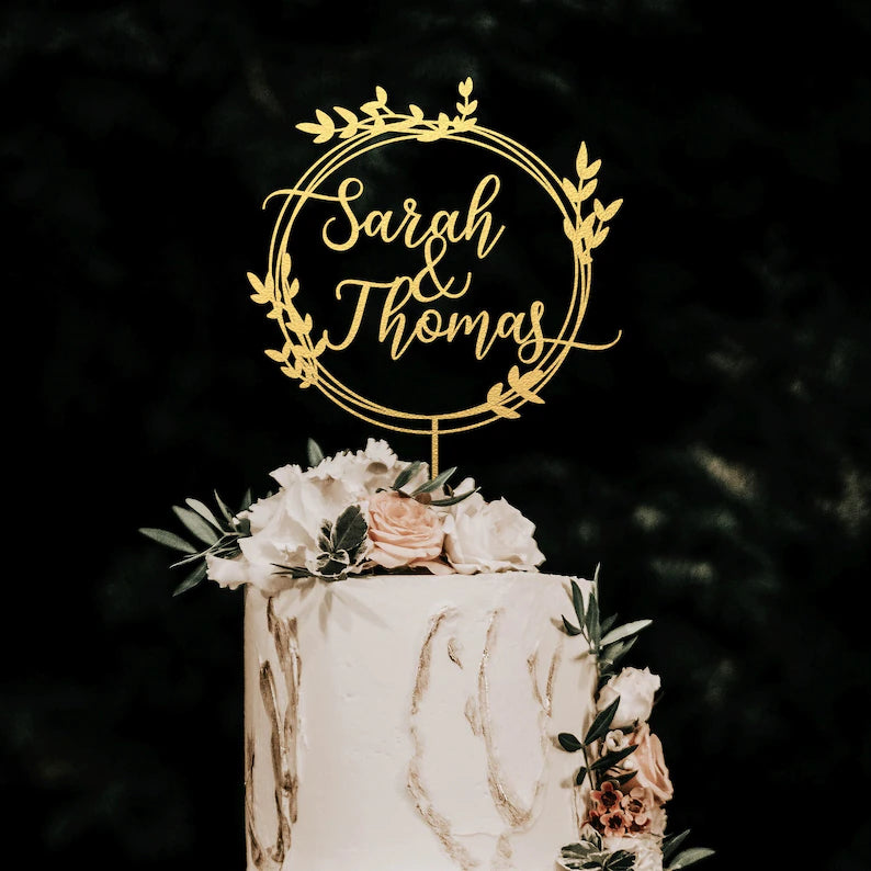 A wedding cake topped with a personalized acrylic cake topper. The topper features the names "Sarah &amp; Thomas" in elegant script, surrounded by a circular design with floral elements. The cake is decorated with white frosting, light pink roses, and greenery, set against a dark background.