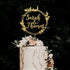 A wedding cake topped with a personalized acrylic cake topper. The topper features the names "Sarah & Thomas" in elegant script, surrounded by a circular design with floral elements. The cake is decorated with white frosting, light pink roses, and greenery, set against a dark background.