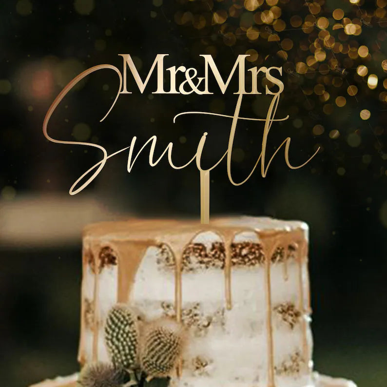 A decorative acrylic cake topper that reads "Mr &amp; Mrs Smith" is displayed on a layered wedding cake. The cake features a smooth white frosting with gold drip decoration. The background is softly blurred with warm bokeh lights, creating a festive atmosphere.