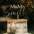 A decorative acrylic cake topper that reads "Mr & Mrs Smith" is displayed on a layered wedding cake. The cake features a smooth white frosting with gold drip decoration. The background is softly blurred with warm bokeh lights, creating a festive atmosphere.