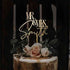 A cake topper featuring the phrase "MR & MRS" in an elegant font, along with the name "Smith." The topper is placed atop a tiered cake adorned with soft pink roses and greenery, including dark leaves. The cake has a gold finish, adding a touch of sophistication to the overall presentation. The background is dark, enhancing the visual impact of the cake and topper.