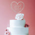 An elegant three-tier white wedding cake sits against a soft pink backdrop. Atop the cake is a personalized acrylic cake topper in a heart shape, featuring the names "Joseph" and "Sarah" along with the date "05.10.2023." The topper is clear and stylishly designed. Adorning the cake are delicate pink flowers, including a large peony at the top and smaller ranunculus flowers on the side, enhancing the romantic aesthetic.