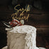 A round wedding cake with a textured gray frosting, topped with a golden acrylic cake topper that reads "Sarah & Richard." The cake is decorated with eucalyptus leaves and small red berries, set against a dark wooden background.