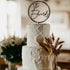 A three-tiered white wedding cake adorned with textured icing. Atop the cake is a black acrylic cake topper featuring the name "The Edwards" inside a circular design. Surrounding the base of the cake are dried wheat-like plants, set against a softly lit background that suggests an elegant event setting.