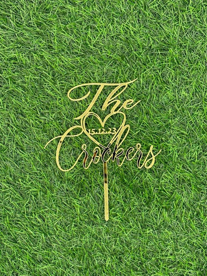 A golden acrylic wedding cake topper is displayed on a green grass background. The topper features the text "The Crockers" with a heart symbol and the date "15.12.23" integrated into the design.