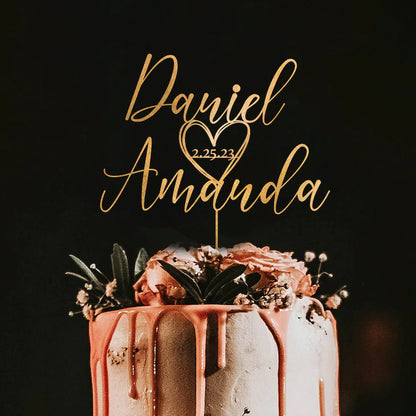 A wedding cake with a personalized acrylic cake topper featuring the names "Daniel" and "Amanda," the date "2.25.23," and a heart design. The cake is decorated with dripping pink frosting and adorned with flowers and greenery on top, set against a dark background.