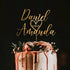 A wedding cake with a personalized acrylic cake topper featuring the names "Daniel" and "Amanda," the date "2.25.23," and a heart design. The cake is decorated with dripping pink frosting and adorned with flowers and greenery on top, set against a dark background.