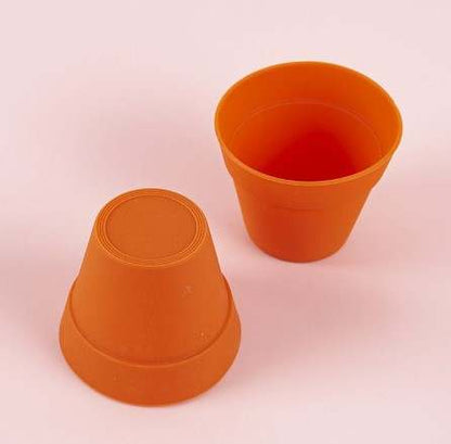 Two orange silicone plant pot molds are shown on a light pink background. One mold is inverted, displaying its base, while the other is right side up, revealing its interior. Both molds have a tapered design typical of plant pots.
