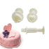 A set of three heart-shaped plunger cutters in white, displayed alongside a pink cake decorated with a heart-shaped topper that says "Valentine" and edible flowers. The cutters consist of two heart shapes and a plunger mechanism, showcasing their use for creating heart-shaped decorations. The cake features a textured surface with small heart designs.