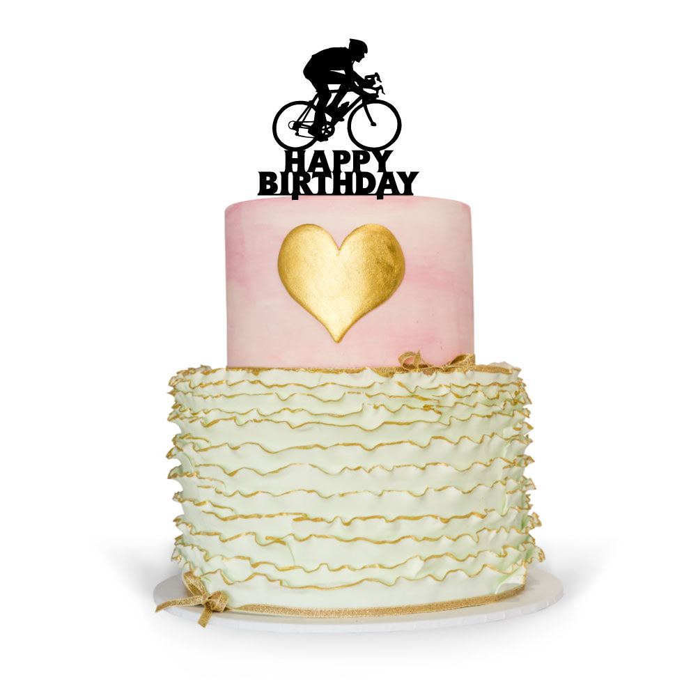 A two-tier birthday cake with a bottom layer featuring green ruffled icing and a top layer in pink with a gold heart decoration. Atop the cake is a black cake topper shaped like a cyclist, alongside the words &quot;HAPPY BIRTHDAY&quot; in bold letters. The cake is presented on a white cake board against a plain background.