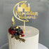 A round acrylic cake topper featuring the words "Ramadan Mubarak" in elegant script, accompanied by a silhouette of a mosque. The topper is gold-colored and is placed on top of a white frosted cake, decorated with a colorful assortment of berries, including raspberries and blueberries, along with golden embellishments and green leaves. The cake has a smooth finish and a dripping icing effect on the sides. The background is softly blurred.