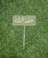 A clear acrylic cake topper with a rectangular shape, featuring elegant gold lettering that reads in Arabic. It is mounted on a thin stick for easy placement on a cake. The topper is set against a backdrop of artificial green grass.
