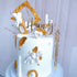 A beautifully decorated white cake featuring a variety of ornate accents. The cake is topped with an acrylic "Happy" and "ANNIVERSARY" topper in a golden script. Surrounding the toppers are decorative elements, including a golden frame, small white cherubs, a heart, and golden leaves. The cake has a smooth white frosting with scattered golden details, giving it an elegant and festive appearance. The overall design suggests a celebration, perfect for anniversaries.