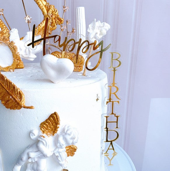 A white cake topped with decorative elements for a birthday celebration. The centerpiece features a gold "Happy" and a golden "BIRTHDAY" sign decorated with embellishments like white flowers, a heart, and a cherub figure. The cake has a smooth finish and is adorned with golden accents.