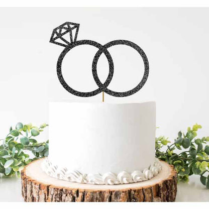 A round white cake is centered on a rustic wooden cake stand. Topping the cake is a black glittery cake topper featuring two interlocking rings, with one ring adorned by a diamond-shaped design. The decorated cake is surrounded by green foliage, adding a touch of color to the presentation.