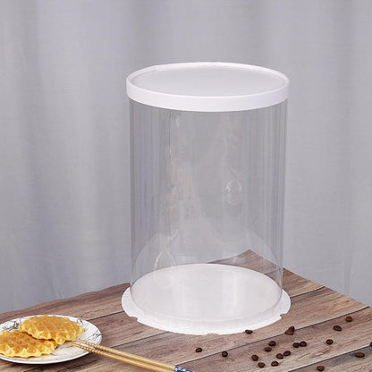 A clear, round PVC cake box with a white lid, measuring approximately 11.8 inches in diameter and 17.7 inches in height. The box has smooth, transparent walls that allow visibility of items inside. It is placed on a wooden surface, alongside a plate of golden, waffle-like pastries and some scattered coffee beans. The background is a soft gray, enhancing the visibility of the cake box and surrounding items.