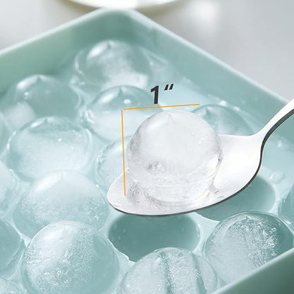 A round ice cube tray filled with spherical ice cubes. One ice cube is being lifted with a spoon, showing its size of approximately 1 inch. The tray is light blue, and the ice cubes are clear and slightly translucent.