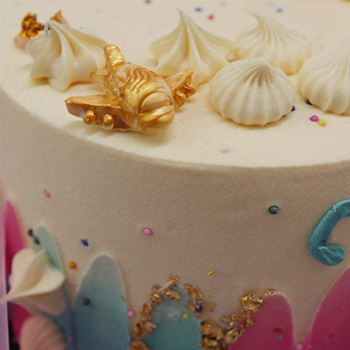 A close-up of a decorated cake featuring a golden airplane decoration on top, surrounded by several swirls of cream and colorful sprinkles. The cake has a pastel-colored border with elaborate designs, showcasing a playful and celebratory theme.