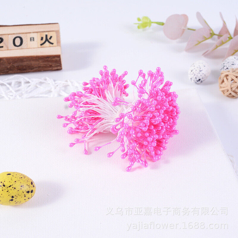 A cluster of pink flower stamens is displayed on a white fabric surface. The stamens consist of multiple thin white threads with tiny pink bead-like tips, arranged to resemble a flower&