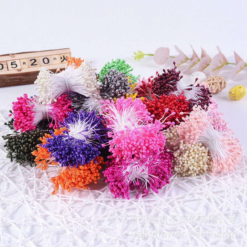 A collection of colorful flower stamens displayed on a textured white surface. The stamens are grouped in clusters, showcasing various colors including pink, purple, white, orange, red, green, and black. Wooden calendar blocks are visible in the background, showing the date, along with decorative elements like dried leaves and a small egg.