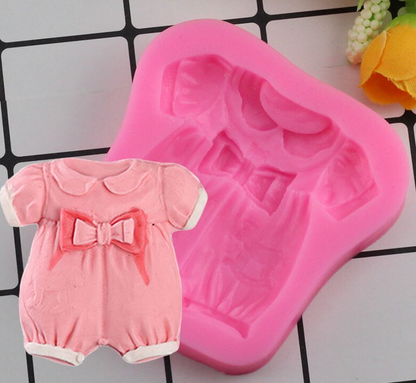 An image of a pink silicone mold shaped like a baby girl dress. The mold features intricate details, including a collar, sleeves, and a bow at the waist. Next to the mold is a small, completed pink dress-shaped figure, showcasing the design as it appears when removed from the mold. The background consists of a grid pattern, with a floral accent in the corner.