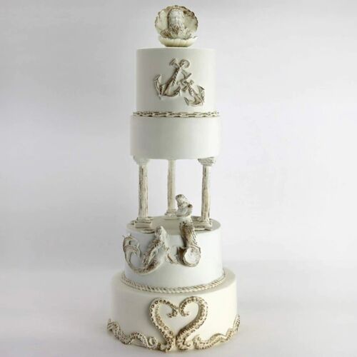 The image features a beautifully designed white cake with multiple tiers. Each tier is adorned with decorative maritime elements, including anchors and seashells. The cake includes intricate detailing with rope-like patterns and mermaid figures. The top tier is crowned with a seashell decoration. The overall aesthetic is elegant and suitable for a nautical-themed celebration.