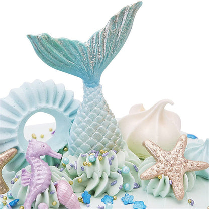 A decorative cake featuring various ocean-themed elements, including a blue and glittery mermaid tail, a light blue shell-shaped piece, a purple seahorse, a beige starfish, and soft swirls of aqua-colored icing. The design incorporates colorful edible decorations like small beads and star-shaped sprinkles.